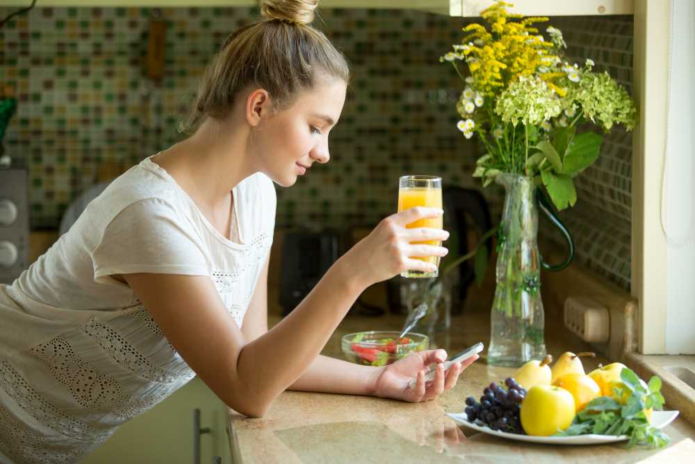 Žena, která je oblečená ve sportovním, pije džus a má kolem sebe ovoce na detoxikaci
